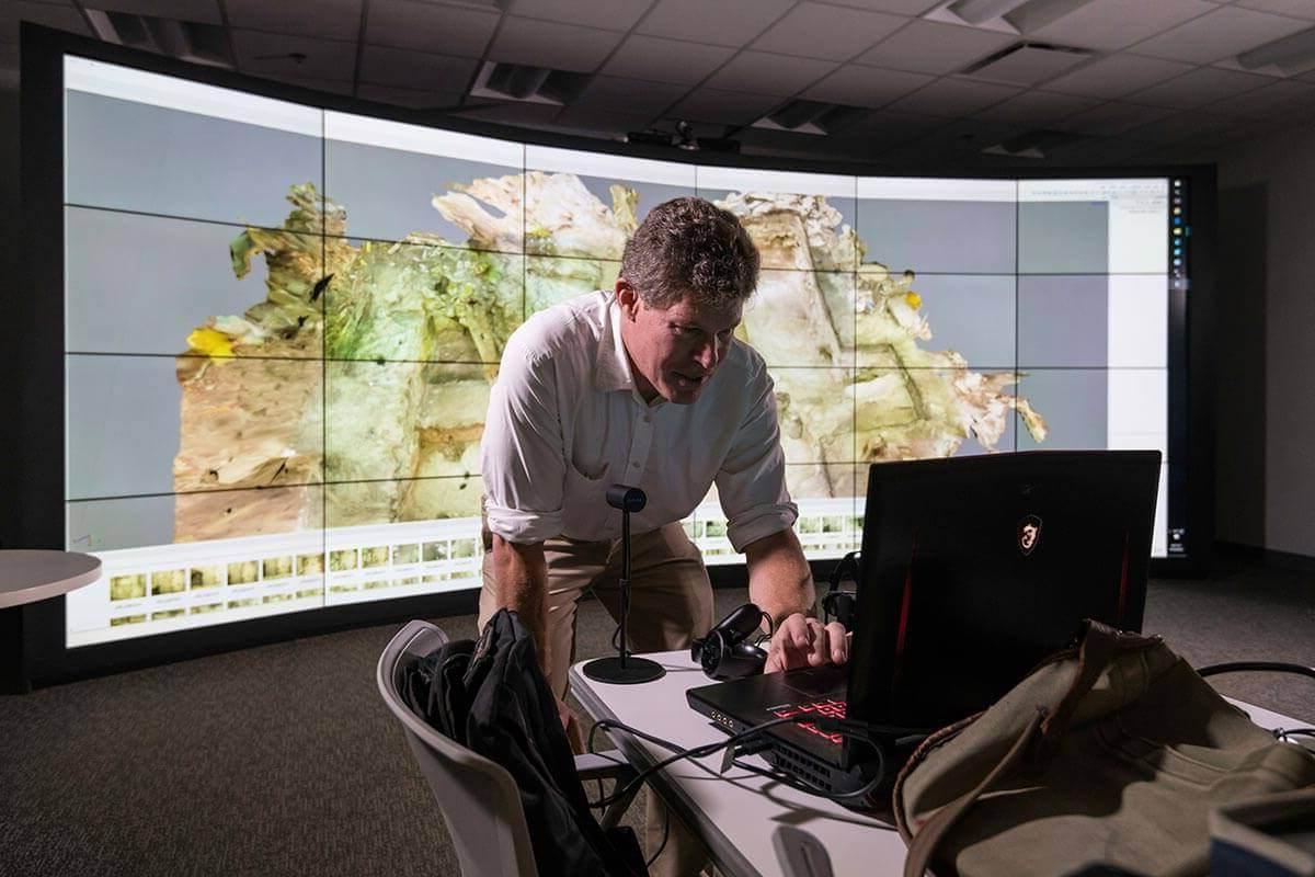 Man leaning over laptop with curved wall monitor display in background at University of Rochester